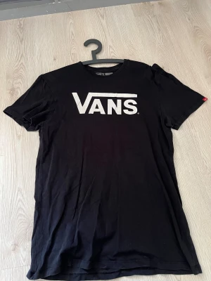 Svart Vans t-shirt med logga - Svart t-shirt från Vans i storlek Large med klassisk vit Vans-logga tryckt på bröstet. Modellen har rund hals och korta ärmar, perfekt för en avslappnad streetstil. Materialet är mjuk bomull som känns skönt mot huden.