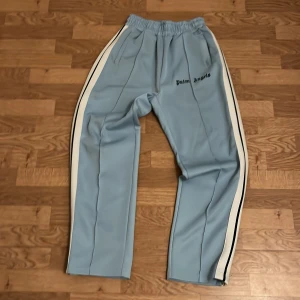 Palm Angels ljusblå trackpants - Snygga ljusblå trackpants från Palm Angels med vita och svarta sidoremsor. Byxorna har elastisk midja och logotypen broderad på framsidan. Perfekt för en chill och sportig look.