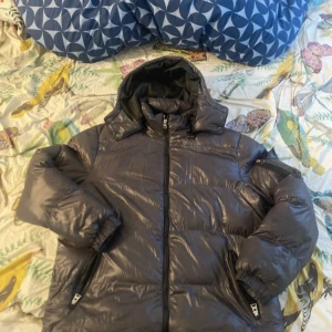 Mörkgrå pufferjacka med huva - Säljer en mörkgrå pufferjacka från Zon i storlek L. Jackan har en stor huva, dragkedja framtill och två sidofickor med dragkedja. Materialet är glansigt syntet och jackan är quiltad för extra värme. Perfekt för kalla dagar och har en schysst oversized vibe.