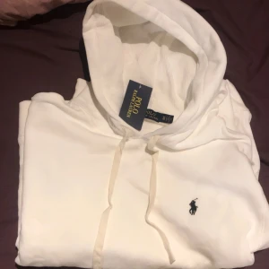 Vit hoodie från Polo Ralph Lauren - Vit hoodie från Polo Ralph Lauren med klassisk broderad logga på bröstet. Tröjan har huva med snörning och är tillverkad i mjuk bomull. Perfekt för en clean och stilren look.