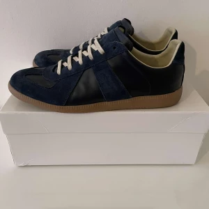 Mörkblå sneakers i mocka och skinn - Snygga mörkblå sneakers från Maison margiela  med ovandel i mocka och skinn, vita snören och klassisk gummisula. Skorna har en stilren design med rund tå och diskreta detaljer, perfekta för dig som gillar en clean och tidlös look.