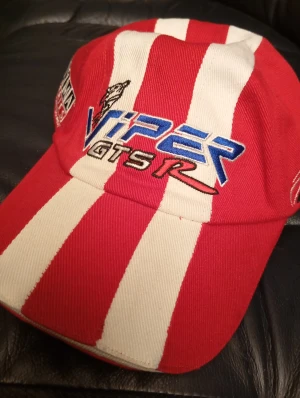 Rödvit Viper GTS-R keps - Snygg keps med breda röda och vita ränder, broderad Viper GTS-R logga i blått, svart och rött framtill samt Optima Batteries patch på sidan. Kepsen har klassisk böjd skärm och är tillverkad i bomull. Perfekt för dig som gillar racing och coola accessoarer.