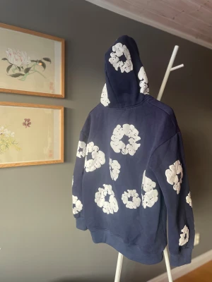 DENIM TEARS - Snygg mörkblå hoodie med stora vita blommönster över hela tröjan. Tröjan har huva, magficka och långa ärmar. Perfekt för dig som gillar streetwear och vill sticka ut med ett coolt mönster. Materialet känns som mjuk bomull och passformen är normal.
