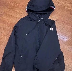 Svart vindjacka från Moncler - Svart vindjacka från Moncler med huva och snörning, dragkedja framtill och Moncler-logga på ärmen. Snygg detalj med röd och vit rand på insidan av huvan. Jackan har fickor med knappstängning och är tillverkad i lätt polyester.