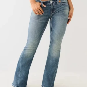 True religion blå bootcut jeans med broderade fickor - Snygga blå jeans från True Religion med bootcut passform och låg midja. Jeansen har markerade sömmar och broderade bakfickor med knappar. Materialet är klassisk jeans och färgen är ljusblå med slitningar framtill och baktill.