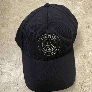 Svart PSG keps med broderad logga - Snygg svart keps från Paris Saint-Germain med broderad logga framtill och texten 'ICI C'EST PARIS' på insidan. Justerbar rem baktill för perfekt passform. Kepsen är tillverkad i slitstark polyester och har klassisk böjd skärm.