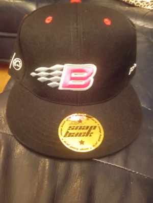 Svart snapback keps Lynx B Racing - Svart snapback keps med broderad B Racing-logga i rött och vitt framtill, samt Lynx och BRP tryck på sidan. Platt skärm med guldfärgad snapback-sticker. Ventilationshål med röda detaljer. Perfekt för dig som gillar motorsport och streetstyle.