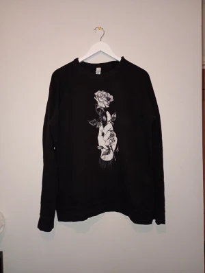 Svart sweatshirt med rosprint, storlek L - Svart sweatshirt i storlek L med en snygg vit print på bröstet som föreställer en hand som håller en ros. Tröjan har rund hals och långa ärmar. Materialet är mjukt och består av 80% bomull, 10% polyester och 10% återvunnen polyester. Perfekt för dig som gillar coola grafiska tryck.