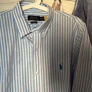 Blårandig skjorta från Polo Ralph Lauren - Snygg klassisk skjorta från Polo Ralph Lauren i blått och vitt med vertikala ränder. Classic fit med button-down krage och broderad logga på bröstet. Skjortan har långa ärmar och är gjord i mjuk bomull. Perfekt för dig som gillar stilrena och fräscha plagg.