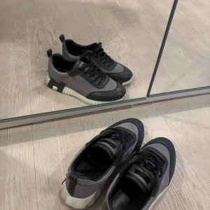 Grå och svarta sneakers med mockadetaljer - Säljer ett par snygga sneakers i grått och svart med coola detaljer på sidorna och vit räfflad sula. Ovandelen är i mocka och skinn, med svarta snören och rund tå. Perfekt för dig som gillar stilrena och moderna skor.