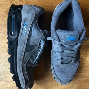 Nike Air Max 90 grå sneakers - Nike Air Max 90 sneakers i grå mocka och mesh med svarta detaljer och blå logga. Klassisk chunky sula med synlig Air Max-enhet och snörning framtill. Snygg streetstil med coola materialmixar och tydlig Nike branding på häl och plös.