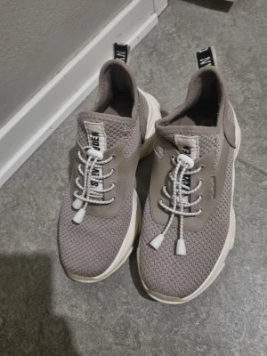 Beige sneakers från Steve Madden - Snygga chunky sneakers från Steve Madden i beige och grå toner. Ovandelen är stickad mesh med detaljer i syntet och coola snörningar med vita snören. Platt, tjock sula i vitt och beige. Perfekt för dig som gillar streetstyle och vill ha bekväma skor med trendig look.