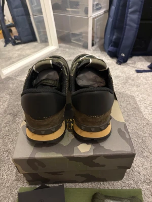 Valentino Rockrunner - Snygga Valentino Garavani Rockrunner sneakers i camouflage-mönster med gröna, beige och svarta toner. Ovandel i mesh och mocka, platt sula med pyramidformade detaljer och rund tå. Klassisk snörning och logga på plösen. Perfekt för dig som gillar streetstyle och exklusiva märken.