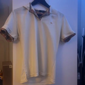 Beige pikétröja från Burberry - Säljer en stilren vit pikétröja från Burberry med klassiskt rutigt mönster på kragen och ärmsluten. Tröjan har en broderad logotyp i gult på bröstet och korta ärmar. Perfekt för en elegant och avslappnad look. Vet dock inte om det är en förfalskning. ‼️Den är inte XL i man storlek utan i barn storlek. Passar nog 150-160 ‼️