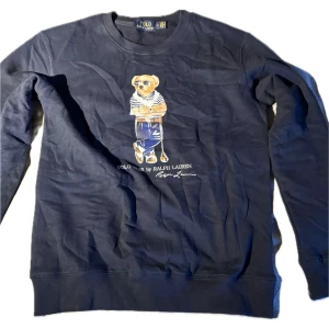 Mörkblå långärmad tröja Polo Bear - Säljer en mörkblå långärmad tröja från Polo Ralph Lauren med ikoniska Polo Bear-trycket på bröstet. Tröjan har rund hals och ribbade muddar. Materialet är mjuk bomull och passformen är normal. Perfekt för dig som gillar klassisk streetwear med en twist.