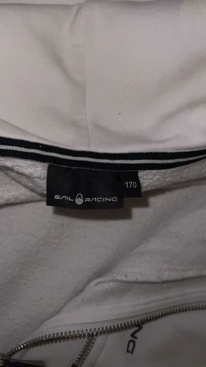 Vit hoodie med dragkedja från Sail Racing - Vit hoodie från Sail Racing med hel dragkedja framtill och snörning i huvan. Tröjan har en bröstficka med dragkedja och logotypen broderad på vänster sida. Mjuk insida och ribbade muddar vid ärmslut och nederkant. Perfekt för chill dagar.