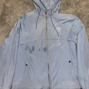 Ljusblå vindjacka från Trapstar - Snygg ljusblå vindjacka från Trapstar med huva, dragkedja med T-detalj och logga på bröstet. Jackan har snörning i huvan, två fickor med knappstängning och är tillverkad i glansig polyester. Perfekt för dig som vill ha en clean och stilren look.