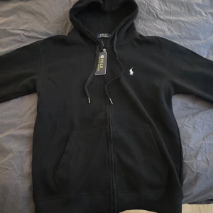 Svart hoodie från Polo Ralph Lauren - Svart hoodie från Polo Ralph Lauren med klassisk vit logobrodyr på bröstet.  Helt ny med taggar Endast provad Lite mindre i storleken