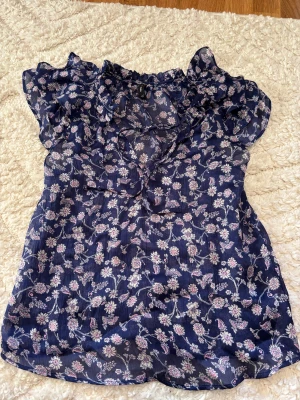 Blommig blå blus från mango  - Supersöt blommig blus från MNG i mörkblått med ljusrosa och vita blommor. Blusen har volangdetaljer vid halsen och är kortärmad med lätt transparent material. Säljer då den blivit för liten. 