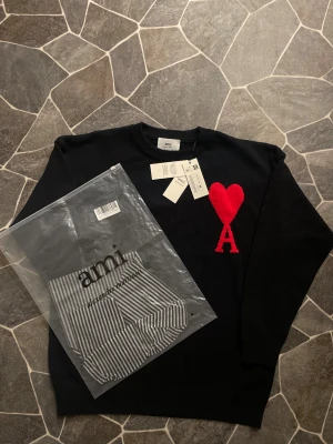 Svart stickad tröja med rött hjärta från Ami - Svart stickad tröja från Ami Alexandre Mattiussi med ett stort rött hjärta och bokstaven A broderat på bröstet. Rund hals och ribbade muddar. Tröjan är i normal passform och tillverkad av mjuk bomull. Perfekt statement-plagg för dig som gillar stilrena detaljer.