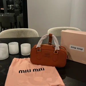 Brun handväska från Miu Miu - Snygg brun handväska från Miu Miu i skinn med guldfärgade detaljer och dragkedja. Väskan har ett stilrent, rektangulärt format och kommer med original dustbag och box. Perfekt storlek för det viktigaste och har ett innerfack med logotyp.