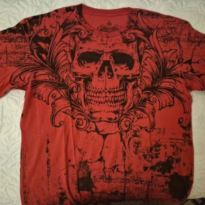 Röd Archaic t-shirt med dödskalletryck, Jesse Pinkman core - Säljer en röd t-shirt från Archaic i storlek XL med ett stort svart dödskalletryck och detaljerade ornament på framsidan. Baksidan har sprucket mönster och logga. Materialet är 100% bomull och passformen är normal. Perfekt för dig som gillar edgy streetstyle.