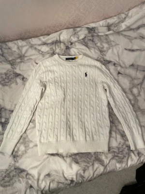 Vit kabelstickad tröja Polo Ralph Lauren - Vit kabelstickad tröja från Polo Ralph Lauren med klassisk rund hals och broderad marinblå logga på bröstet. Tröjan har långärmad passform och ribbade muddar vid ärmslut och nederkant. Perfekt för dig som gillar stilrena och tidlösa plagg.