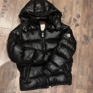 Svart Moncler mata pufferjacka  - Svart pufferjacka från Moncler med glansig finish, huva och dragkedja framtill. Jackan har Moncler-logga på ärmen och coolt serietidningsmärke på insidan. Flera fickor med dragkedja och ribbade muddar. Perfekt för dig som vill ha en snygg och varm jacka.