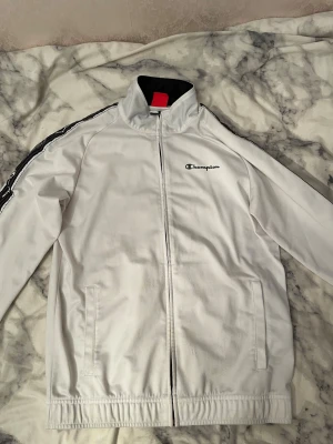 Vit vindjacka från Champion M - Snygg vit vindjacka från Champion i storlek medium. Jackan har svarta detaljer längs ärmarna med Champion-loggor, hög krage och dragkedja framtill. Tillverkad i polyester och har två fickor på sidorna. Perfekt för en sportig och clean look.