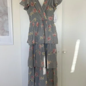 Blommig långklänning med volanger - Säljer en långklänning i ljusgrått med blommigt mönster i rosa och gult. Klänningen har volanger i flera lager, omlottliv med v-ringning, korta volang ärmar, i ryggen så har den band så man kan knyta. Den är aldrig använd och prislappen sitter fortfarande kvar!!✨🩷