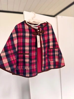 Rutig cape från Rockin' Baby, rosa/blå - Superfin cape från Rockin' Baby i rutigt mönster med rosa, blå och vita toner. Jackan har bruna kanter, två fickor fram och knytning vid halsen. Utsidan är i mjuk ullmix och insidan är fodrad i rosa bomullstyg. 