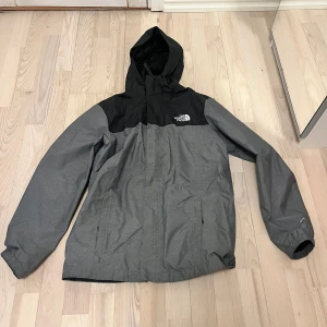 Grå och svart jacka från The North Face - Säljer en grå och svart jacka från The North Face med huva och dragkedja. Jackan har en stilren design med långärmade ärmar och är perfekt för kyligare väder.