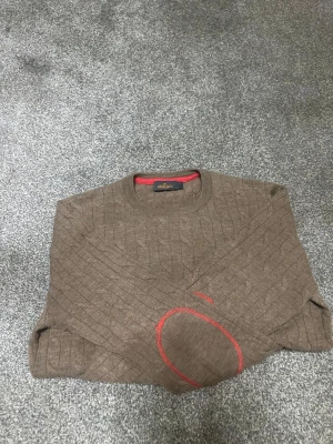 Brun kabelstickad tröja Morris - Brun kabelstickad tröja från Morris med rund hals och snygga röda detaljer, bland annat ett broderat emblem på bröstet och röda armbågslappar. Tröjan har en klassisk look och är perfekt för dig som gillar stilrena plagg med lite edge.