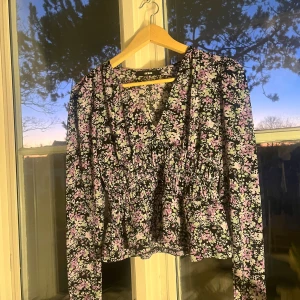Blommig blus med v-ringning från Bikbok - Säljer en blommig blus från Bikbok i storlek S. Blusen har v-ringning, långärmade ärmar och smockad midja som ger en snygg siluett. Färgerna är svart, lila och vit med ett blommigt mönster. Perfekt för dig som gillar romantiska vibes och detaljer.