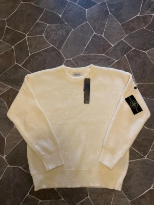 Vit stickad tröja från Stone Island - Vit stickad tröja från Stone Island med klassisk rund hals och ribbade muddar. Tröjan har den ikoniska Stone Island-patchen på vänster ärm och är tillverkad i mjukt material som ger en clean och stilren look. Perfekt för dig som gillar minimalistisk stil. Passar både M och L!