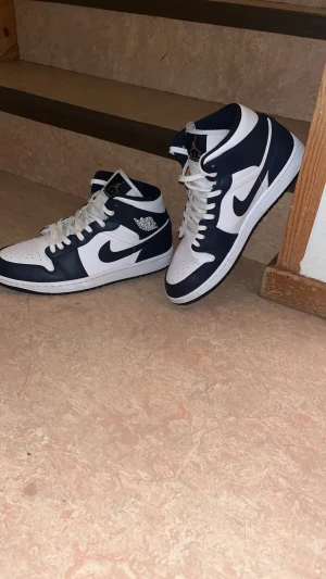 Nike Air Jordan 1 Mid marinblå/vit - Säljer ett par Nike Air Jordan 1 Mid sneakers i marinblått och vitt. Skorna har klassisk snörning, rund tå och platt sula. Ikonisk Jordan-logga på sidan och Nike Swoosh i svart. Tillverkade i skinn med snygg kontrast mellan färgerna. Perfekt för dig som gillar streetwear.