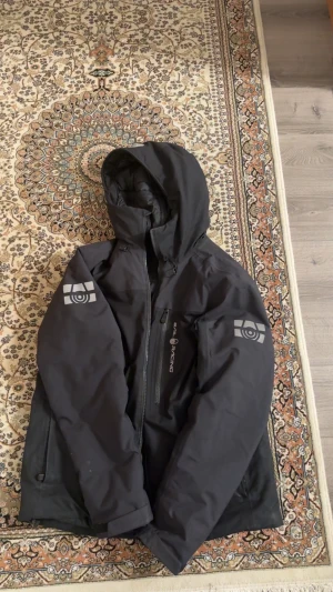 Svart pufferjacka från Sail Racing - Svart vinterjacka från Sail Racing med huva och coola loggor på båda ärmarna. Jackan har dragkedja framtill och flera praktiska fickor. Perfekt för dig som vill ha en stilren och funktionell jacka med sportig vibe.