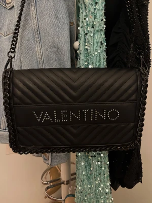 Svart quiltad axelväska Valentino - Snygg svart axelväska från Valentino med kedjerem och quiltad design. Väskan har ett stort VALENTINO-tryck med nitar på framsidan och insida i rött med praktisk dragkedja. Perfekt för dig som vill ha en statementväska med lyxig känsla. Ett år gammal knappt använd💓