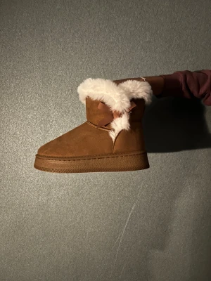 Bruna boots med vit päls - Mysiga bruna boots med vit päls runt kanten och en söt rosett på sidan. Skorna har platt sula och är tillverkade i mocka material, perfekta för kalla dagar. Stilen är enkel men ändå trendig och passar dig som gillar att hålla värmen med stil. Aldrig använda. Hör av dig vid minsta fundering.