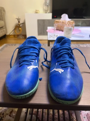 Blåa Puma fotbollsskor med snörning - Säljer ett par blåa Puma fotbollsskor med vit logga och coolt mönster på sidorna. Skorna har snörning och en mintgrön sula som ger en snygg kontrast. Ovandelen är i syntetmaterial och designen är sportig och modern, perfekt för dig som vill sticka ut på planen.