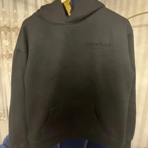 Svart Essentials Fear of God hoodie - Svart hoodie från Essentials Fear of God med diskret logga på bröstet och stor logotyp på ryggen. Tröjan har huva, känguruficka och lång ärm. Perfekt för dig som gillar streetwear och vill ha en clean look.
