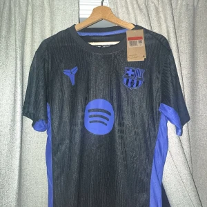 FC Barcelona svart/blå fotbollströja - Snygg FC Barcelona fotbollströja i svart med blå detaljer och logga. Tröjan har korta ärmar, rund hals och är tillverkad i lätt polyester som andas. Spotify-logga på bröstet och klubbmärke på vänster sida. Perfekt för dig som vill sticka ut på planen. Passar även L