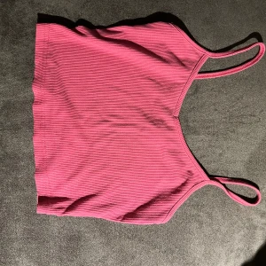 Rosa ribbad linnetopp med smala axelband - Säljer en snygg rosa linnetopp med smala axelband och ribbad struktur. Toppen är croppad och har en enkel, stilren design som är perfekt för varma dagar. Materialet känns mjukt och stretchigt, vilket gör den skön att bära. Passar dig som gillar färg och enkelhet.