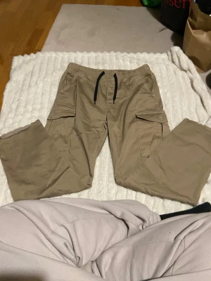 Beiga cargopants med snörning - Säljer ett par beiga cargopants från Vailent, Carlings med svarta snören i midjan och flera fickor med lock och knapp. Byxorna har avslappnad passform och är tillverkade i mjuk bomull, perfekta för chill dagar eller streetwear-stil.