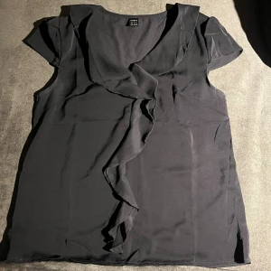 Svart blus med volang från SHEIN - Svart blus från SHEIN i storlek XS. Blusen har korta ärmar och en snygg v-ringad hals med volangdetalj framtill. Materialet är lätt och känns mjukt mot huden, perfekt för dig som gillar stilrena och feminina plagg.