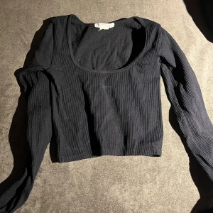 Svart ribbad långärmad topp från Zara - Svart långärmad topp från Zara i ribbad bomull med rundad, djup urringning. Modellen är croppad och har en slim passform som sitter snyggt mot kroppen. Perfekt för dig som gillar stilrena och enkla plagg med lite edge.