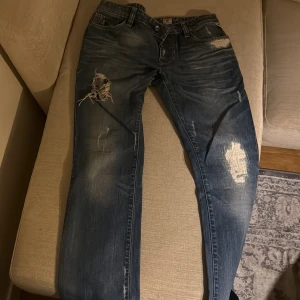 Blå slitna jeans med trasiga detaljer Jack and Jones  - Säljer ett par blå jeans med coola slitningar och trasiga detaljer på benen. Jeansen har klassisk femficksdesign, knapp och dragkedja i gylfen samt en snygg tvättad look. Perfekt för dig som gillar en avslappnad och edgy stil.