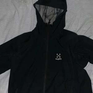 Svart regnjacka från Haglöfs med huva - Svart regnjacka från Haglöfs med hel dragkedja och justerbar huva med meshfoder. Jackan har minimalistisk design och logga på bröstet. Perfekt för blöta dagar och outdoor-äventyr.