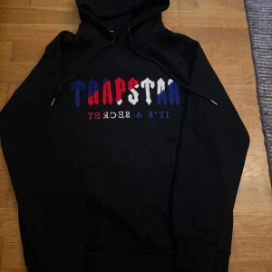 Trapstar hoodie - Svart hoodie från Trapstar med broderad logga i rött, vitt och blått på bröstet. Klassisk huva med snörning och känguruficka framtill. Snygg streetwear-vibe och perfekt för dig som gillar statement-plagg.
