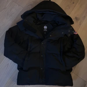Svart dunjacka från Canada Goose - Säljer en svart dunjacka från Canada Goose med huva och klassisk logga på ärmen. Jackan har dragkedja och knappar framtill, är vadderad och perfekt för kalla dagar. Snygg och tidlös modell som håller värmen hela vintern.